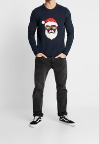 Marineblauer Pullover mit einem Santa-Gesicht-Design, das eine Sonnenbrille, einen roten Hut und weißen Bart zeigt. Kombiniert mit grauen Hosen und schwarzen Turnschuhen.