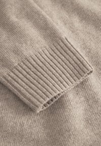 Beige strikket sweater tæt på, med ribbede manchetter, glat tekstur og blødt stof, der fremhæver enkelheden i design og farve.