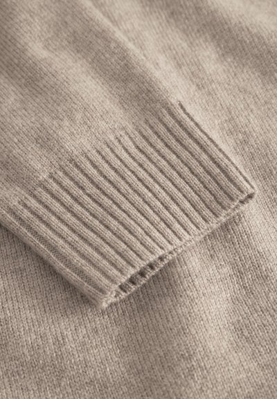 Beige knitted sweater close-up, featuring a ribbed cuff, smooth texture, and soft fabric, highlighting the simplicity in design and color.