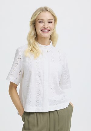 Blond kvinne smilende, iført en hvit kortermet bluse med hullmønster og krage, og olivengrønne bukser, hendene i lommene, på hvit bakgrunn.