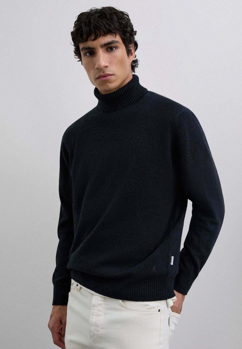 Suéter negro de cuello alto hecho de un material texturizado, con un cuello y puños acanalados, combinado con pantalones blancos.