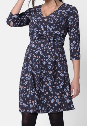 Vrouw draagt een marineblauwe knie-lange bloemenjurk met roze en lichtblauwe bloemen, 3/4 mouwen, V-hals en een gesloten tailleceintuur.