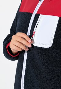 Veste en polaire avec un corps bleu marine, des accents rouges et une poche zippée blanche. Texture douce avec une fermeture éclair contrastante et un cordon de serrage.