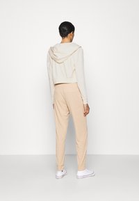 Persona in piedi di spalle, che indossa un hoodie corto bianco, pantaloni a coste beige e sneakers alte bianche, su uno sfondo bianco uniforme.
