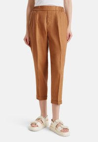 Brązowe spodnie z lnu z domieszką, z elastycznym pasem, o skróconej długości i bocznymi zakładkami, zestawione z białymi, masywnymi sandałami.