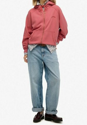 Person trägt eine rote Kapuzenjacke mit Reißverschluss über einem gestreiften Hemd, lockere hellblaue Jeans mit aufgerollten Bündchen und dunkelbraune Loafers vor weißem Hintergrund.