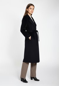 Kvinna står med händerna i fickorna iförd en lång svart bälte kappa, beige byxor och svarta klackboots på vit bakgrund.