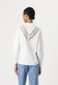 Camicia bianca a maniche lunghe con un'applicazione decorativa di pizzo sulla parte posteriore e colletto classico, abbinata a pantaloni azzurri chiari con texture.