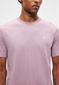 Άνδρας που φοράει απλό κοντομάνικο T-shirt ανοιχτού μοβ χρώματος με μικρό λευκό λογότυπο "M O P" στο αριστερό στήθος, με ορατό λαιμό και χέρια.