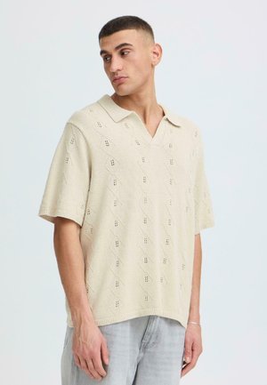 Mann trägt beige kurzärmliges, gestricktes Polohemd mit Rautenmuster, blickt nach links, steht vor hellem Hintergrund.