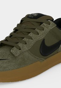Baskets Nike SB vert olive avec un swoosh noir, une semelle marron et des lacets olive, montrant les détails avant et latéraux.