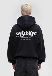 Černá mikina s kapucí s velkým bílým grafickým motivem na zadní straně, obsahující text a loga. Uvolněný střih s tkaničkou na kapuci. Měkká textura látky.