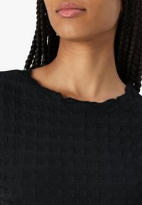 Svart teksturert topp med en bølgete mønster og en scallooped halslinning. Stoffet ser mykt ut med en svak glans.