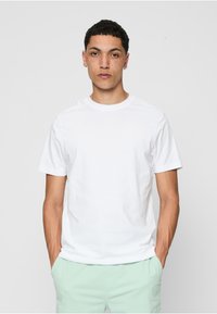 Tânăr cu păr scurt și creț, cercei și pistrui, îmbrăcat cu un tricou alb simplu și pantaloni verzi deschis, cu mâinile în buzunare.