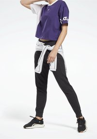 Fialovo-bílé crop top tričko s logem, kombinované s černými tepláky zavázanými v pase. Černé sportovní boty doplňují outfit.