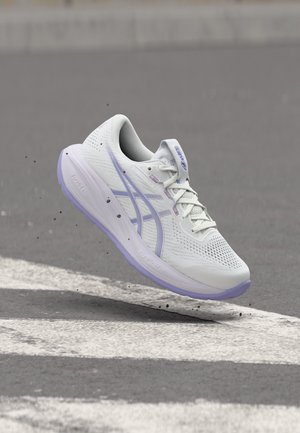 Biało-niebieski but do biegania uniesiony w powietrzu nad asfaltem z białymi oznaczeniami drogowymi, wokół podeszwy unoszą się małe kamyczki.