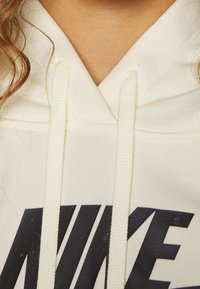 Sweat à capuche blanc avec un col en V, arborant un imprimé noir "NIKE". Dispose de deux cordons de serrage, fabriqué en tissu doux au toucher lisse.