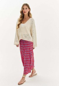 Krémový pletený kardigán s vzormi srdca, skombinovaný s červenou sukňou s modrými abstraktnými tvarmi. Model má na sebe ploché sandále.