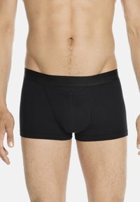 Männlicher Oberkörper, der schwarze, enganliegende Boxershorts mit schwarzem Bund trägt, steht vor einem schlichten weißen Hintergrund.