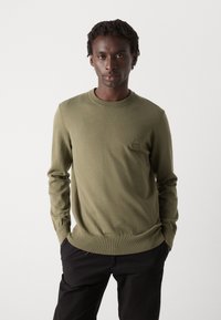 Maglione lavorato a maglia verde oliva con scollo rotondo, polsini a coste e un piccolo logo ricamato sul petto, abbinato a pantaloni neri.