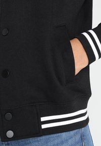 Schwarze Sportjacke mit weißen gestreiften Bündchen, glatter Textur, Druckknöpfen und Seitentaschen, mit tailliertem Design.