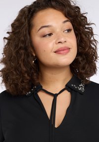 Czarna bluzka z dekoltem w szpic i troczkiem do zawiązania. Kołnierz ozdobiony dekoracyjnymi koralikami i cekinami. Gładka faktura.