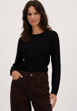 Vrouw met donker golvend haar, gekleed in een zwart geribbeld langemouwen shirt en bruine broek met luipaardprint, hand in de zak, neutrale achtergrond.