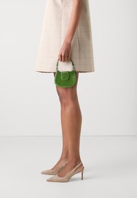 Borsa verde con trama, dalla silhouette curvilinea, dotata di manico superiore e pattina anteriore. Indossata da una persona con una gonna pied de poule beige e scarpe con tacco nudo.