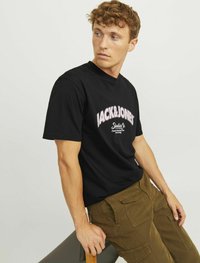Sort bomuld t-shirt med hvid og lyserød "JACK&JONES" logo. Modellen har olivengrønne cargo-bukser på og læner sig op ad en stol. Neutral baggrund.