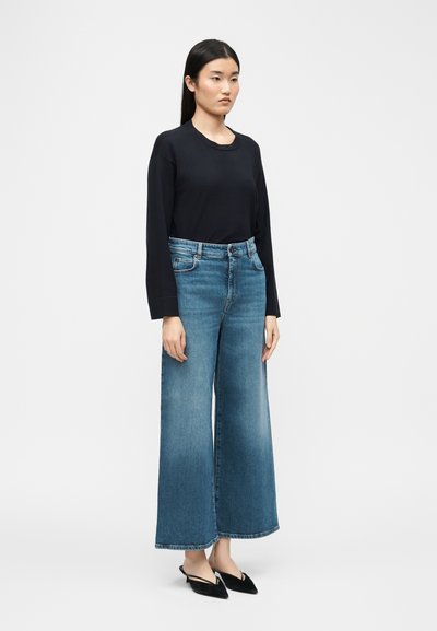 Femme aux cheveux noirs et lisses, portant un haut noir à manches longues, un jean bleu à jambes larges et des mules noires à bout pointu, debout devant un fond uni.