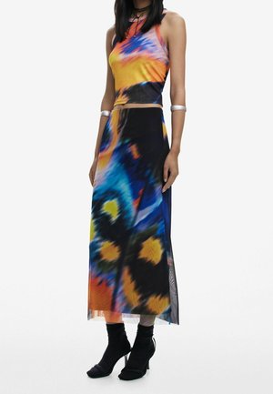 Kvinde iført ærmeløs tie-dye crop top og matchende lang nederdel med abstrakte mønstre, kombineret med sorte hælede sko og sølvarmbånd.