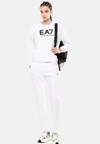 Joven con sudadera y pantalones blancos de EA7 Emporio Armani, llevando una mochila negra sobre un hombro, de pie contra un fondo liso.