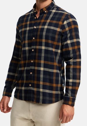 Homme portant une chemise à carreaux bleu marine, marron et beige boutonnée, avec un pantalon beige, debout devant un fond uni.