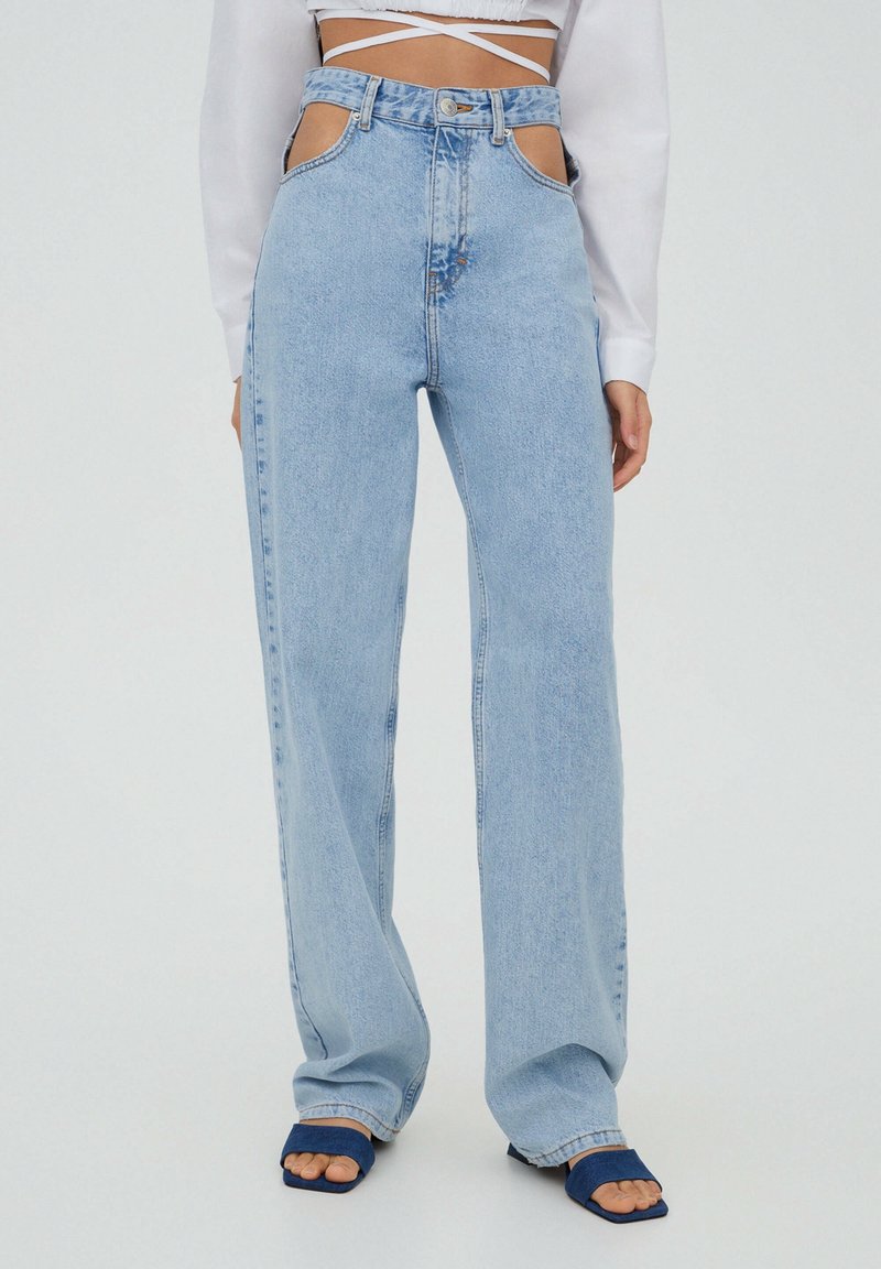 Femme portant un jean taille haute bleu clair avec des découpes sur les côtés, un crop top blanc et des sandales bleues à bouts ouverts sur un fond uni.