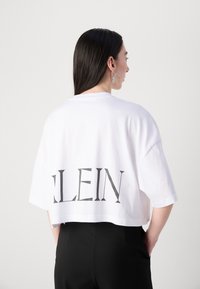 White cropped t-shirt with short sleeves featuring the black text "KLEIN" on the back. Smooth cotton texture, relaxed fit.