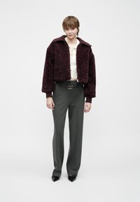 Mladý človek stojí oblečený v textúrovanom tmavobordovom saku, bielom pletenom topu, šedých širokých nohaviciach s opaskom a čiernych špicatých topánkach na jednolatom pozadí.