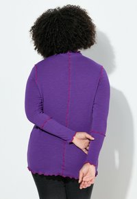 Långärmad lila topp med räfflade röda detaljer längs sömmar och muddar, med avslappnad passform och mjuk textur. Bakifrån vy.