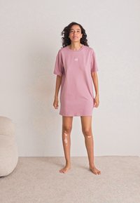 Kvinne med krøllete hår står barbeint på teppet iført en løs rosa t-skjortekjole mot en ensfarget lyse vegg, med en kremfarget sofa i nærheten.
