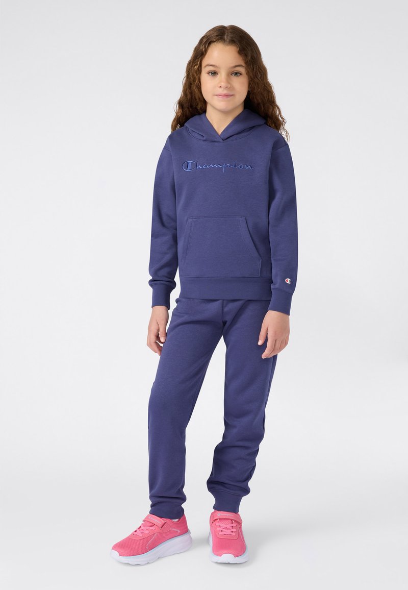 Meisje met krullend haar, gekleed in een blauwe Champion hoodie, bijpassende joggingsbroek en felroze sneakers, staande op een witte achtergrond.