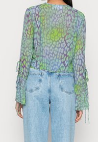 Sheer green and purple patterned blouse with long sleeves and tie details, paired with high-waisted light blue jeans.
