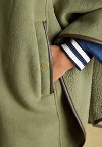 Hand in der Tasche einer olivgrünen Fleecejacke mit braunem Besatz, sichtbarer gestreifter Bündchenabschluss eines blau-weißen Pullovers darunter.