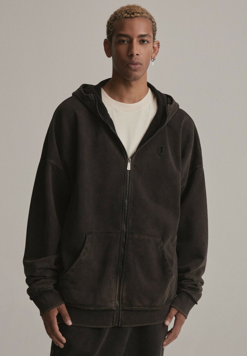 Czarna hoodie na zamek, wykonana z miękkiego materiału, z przednią kieszenią, ściągaczami przy rękawach oraz kapturem. Design ma zużytą teksturę i minimalistyczne logo.