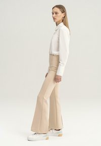 Camisa blanca de manga larga metida dentro de unos pantalones de campana beige de cintura alta, con una textura suave. Zapatillas blancas con detalles en verde claro.