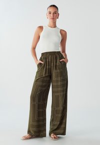 Žena u bijelom bezrukavu rebrastom topu, maslinastim širokim hlačama s geometrijskim uzorcima i bež sandalama, stoji s rukama u džepovima.