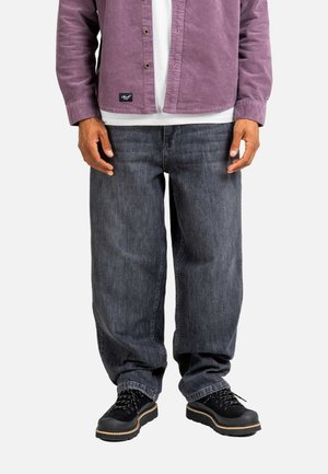 Mann trägt dunkelgraue, lockere Jeans, schwarze Wanderschuhe, ein weißes Hemd und ein lila Hemd mit Knöpfen und steht vor einem weißen Hintergrund.
