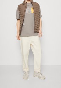 Ruskea pehmustettu liivi harmaan graafisen t-paidan päällä, yhdistettynä kermanvärisiin rentoihin housuihin ja vaaleanharmaisiin lenkkareihin. Yksinkertainen tausta.