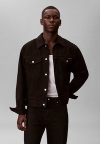 Hombre vistiendo una chaqueta de mezclilla negra sobre una camiseta sin mangas blanca con pantalones negros, de pie frente a un fondo liso de color gris claro.