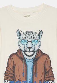 Stylizovaný snežný leopard s modrými slnečnými okuliarmi, modrou košeľou a hnedou zipsovou mikinou na krémovom pozadí.