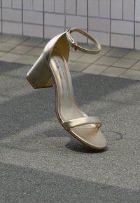 Χρυσό μεταλλικό πέδιλο με χοντρό τακούνι, διαθέτει δύο λεπτά λουράκια και λουρί στον αστράγαλο. Το παπούτσι έχει λεία υφή και στρογγυλεμένη μύτη.