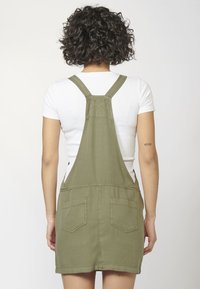 Vestido de tirantes de mezclilla verde oliva con correas ajustables, dos bolsillos traseros y un dobladillo limpio. Combinado con una camiseta blanca ajustada debajo.